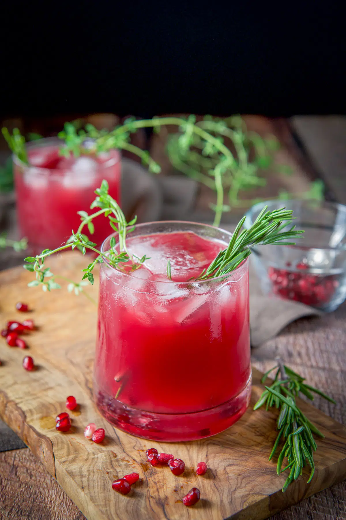 A board with a glass filled with the red drink with arils, rosemary, and thyme in it