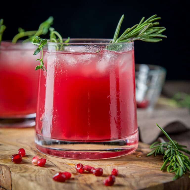 square photo of a glass with the red cocktail with thyme, rosemary, and pomegranate seeds