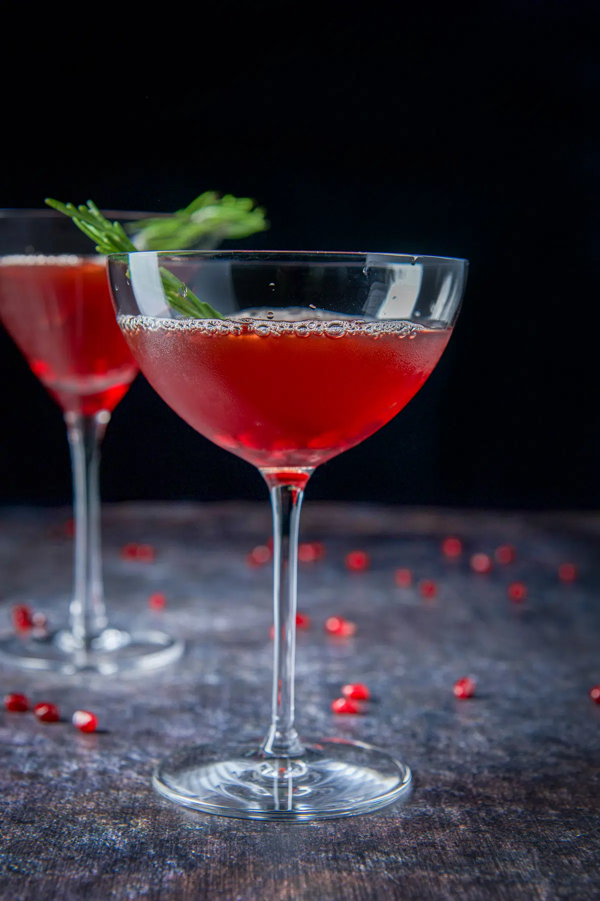 Vertical view of two martini glasses with the cocktail in it with sprigs of rosemary and pomegranate seeds