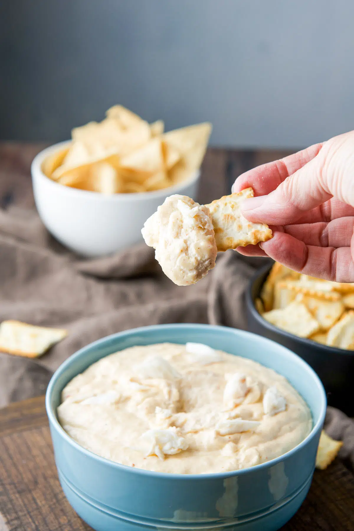 A hand holding a chip with crab dip on it held over the bowl of dip