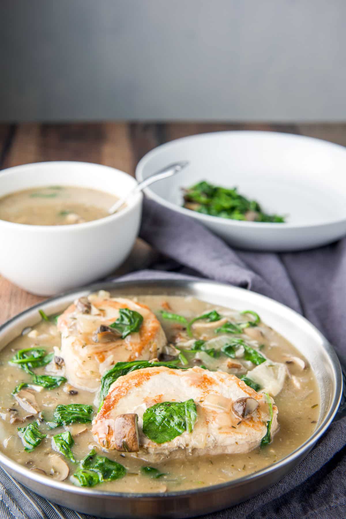 A pan with two pork chops in broth with vegetables in front of a plate and bowl