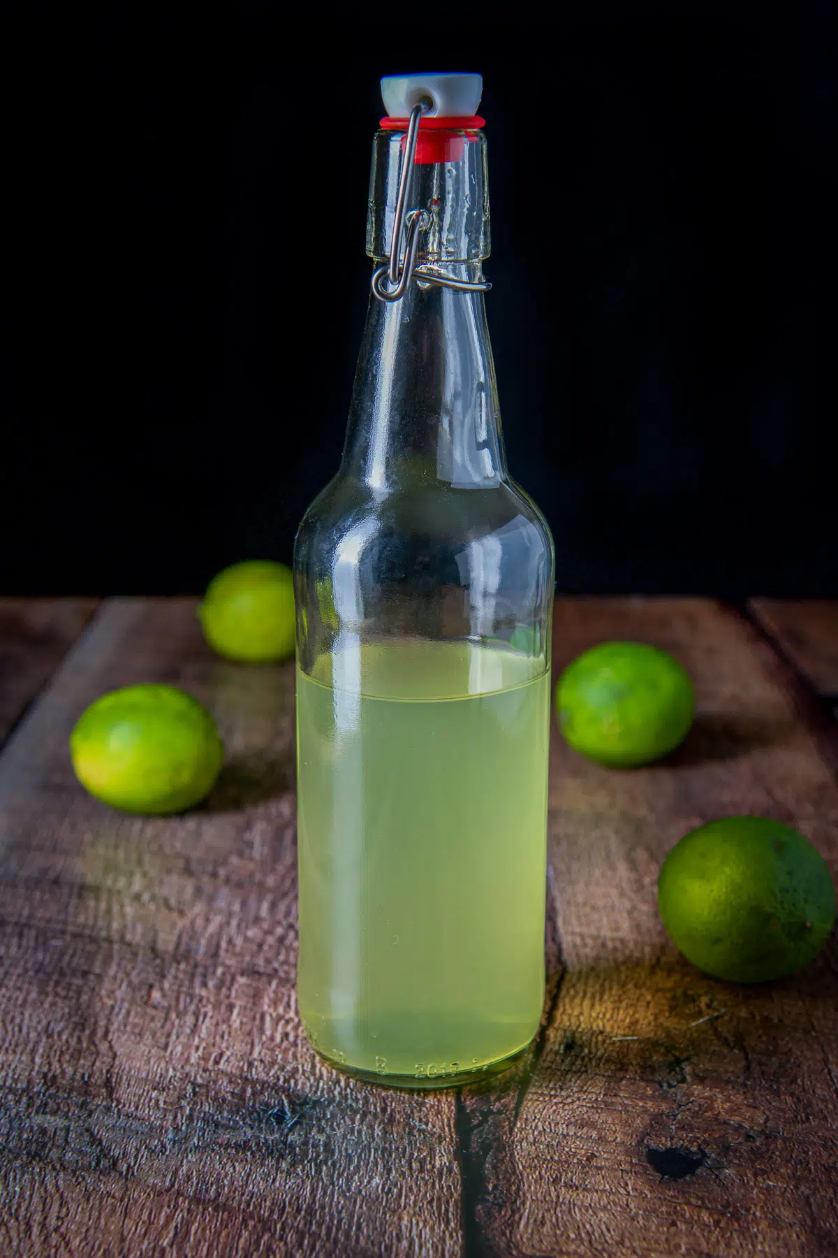 A capped bottle of green vodka with limes in the back