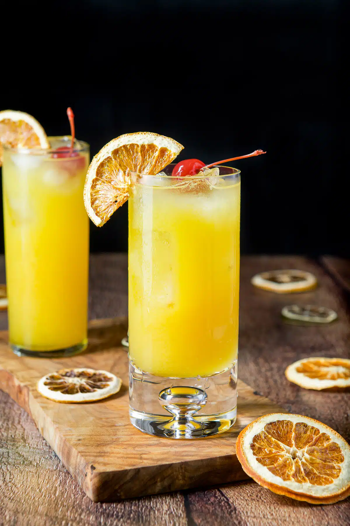 Vertical view of the two glasses on a board with the orange cocktail in it along with orange wheels and cherries