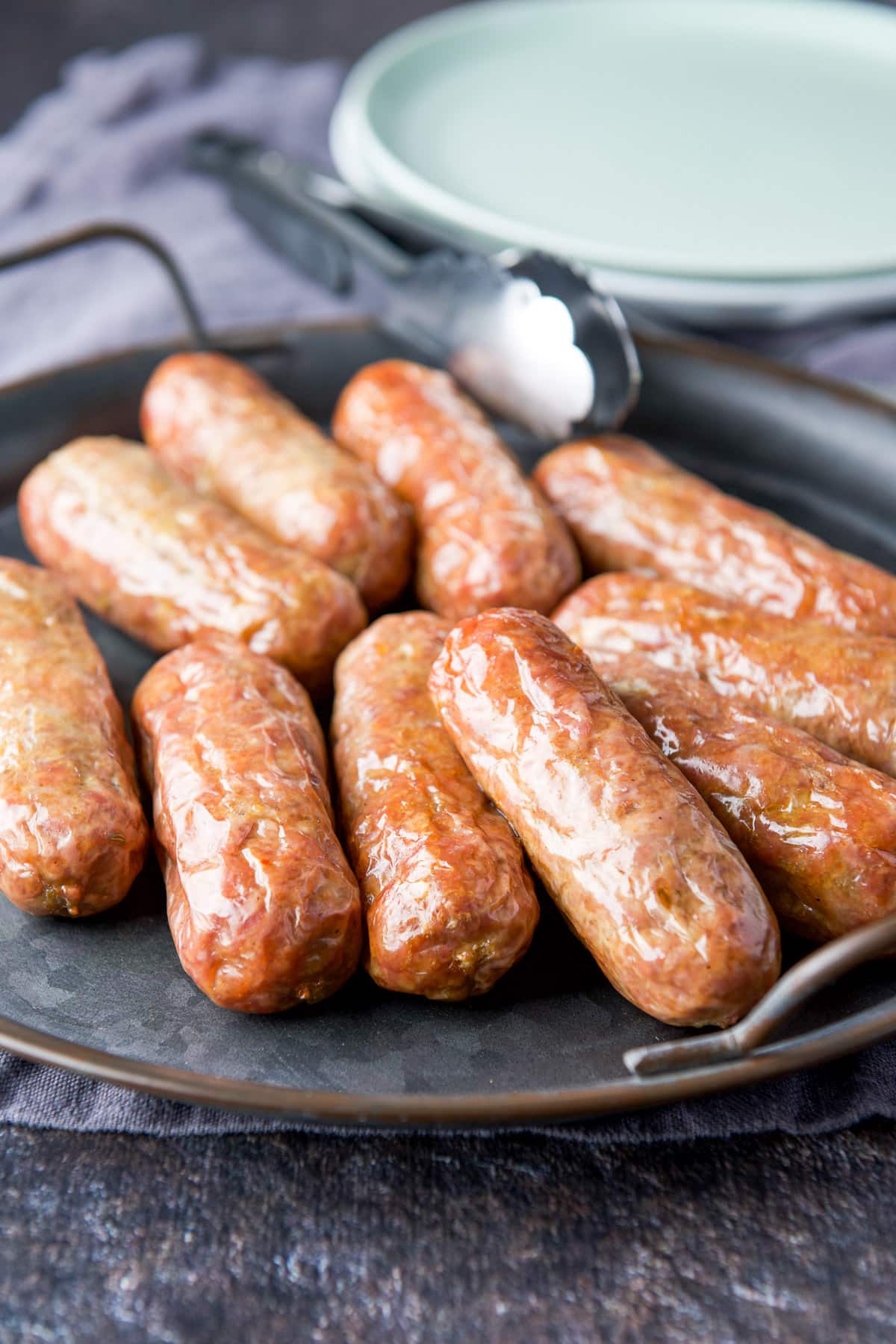 A metal tray filled with sausages, along with tongs and two plates in the back