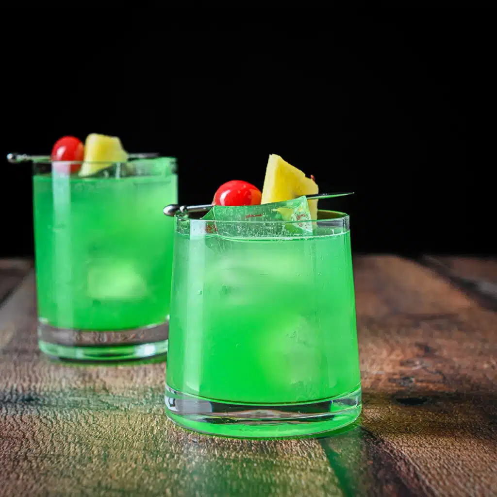 square photo of a green cocktail in two glasses with cherries and pineapple chunks