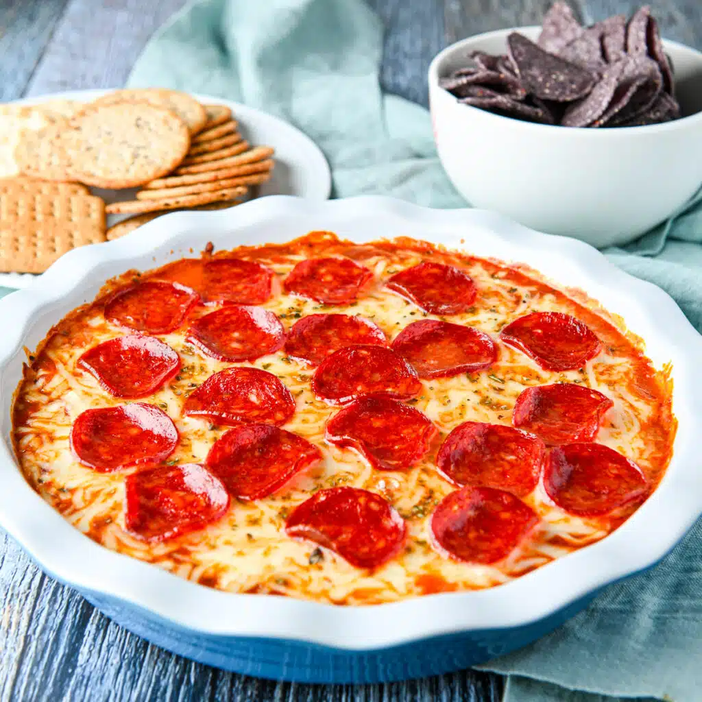 square photo of a pepperoni dip in a pie dish with chips and crackers in the back