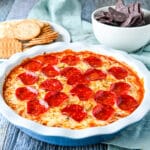 square photo of a pepperoni dip in a pie dish with chips and crackers in the back