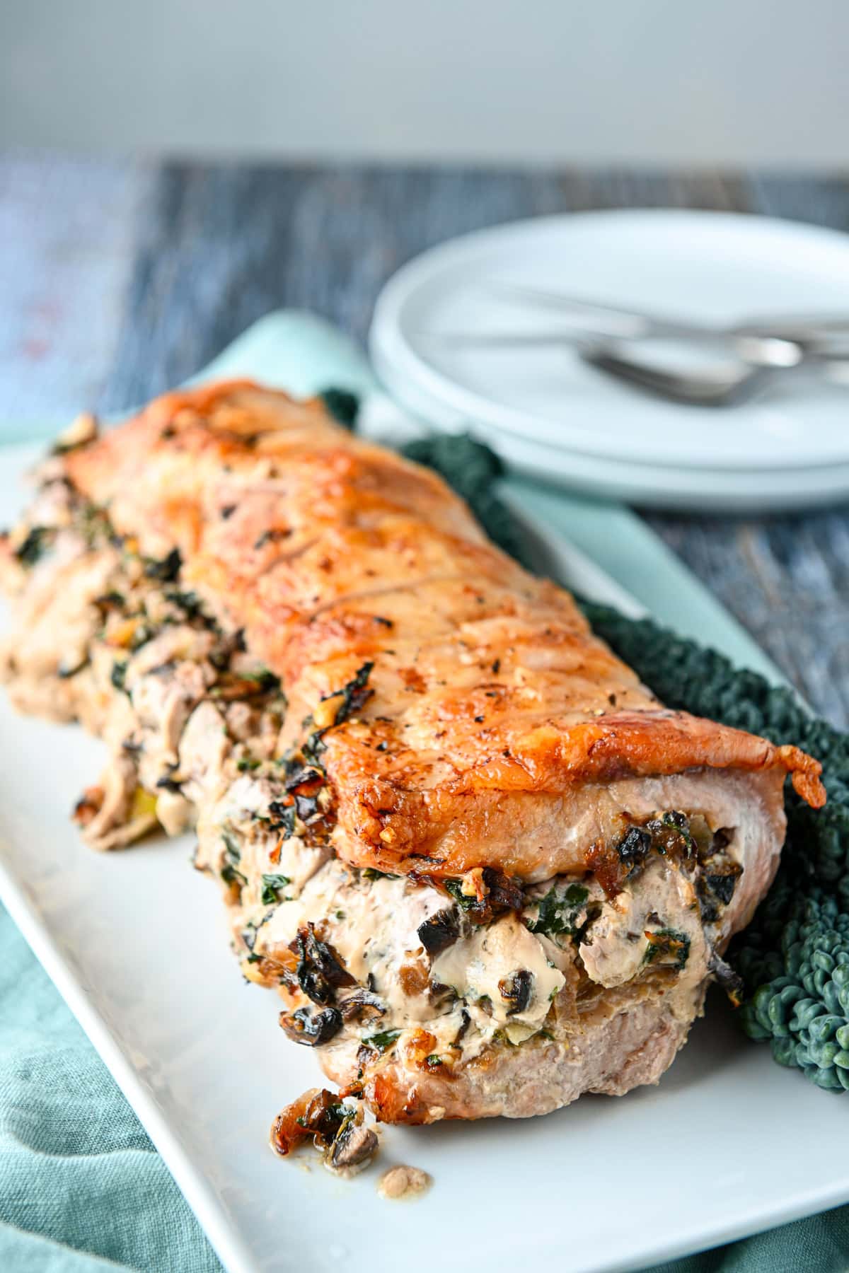 A white rectangular platter with a cooked stuffed loin with white plates in the back