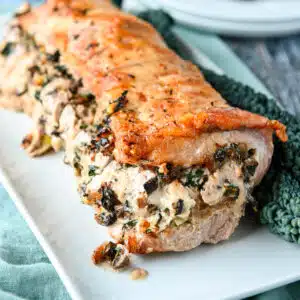 Square photo of a pork loin with stuffing in it on a platter with kale on the side