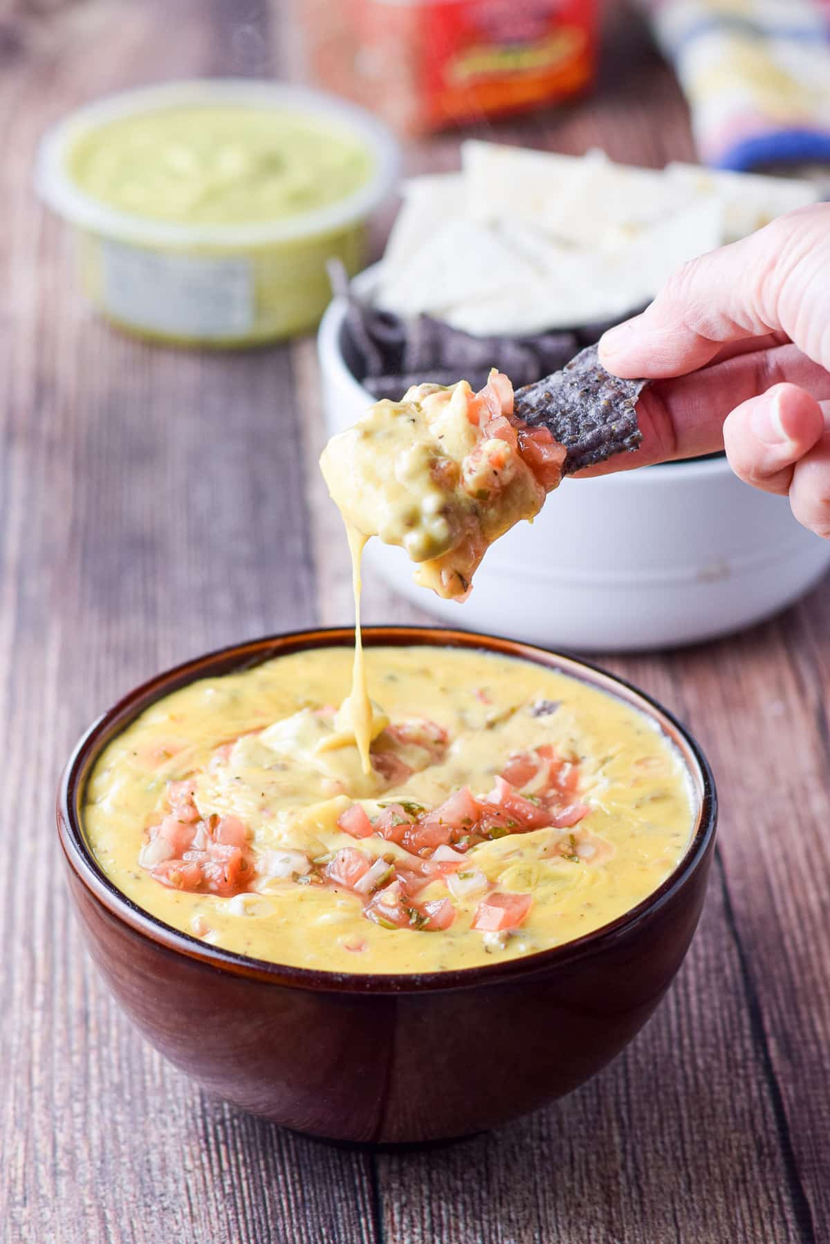 A blue chip in a hand that dipped it in the queso dip with guacamole in the back