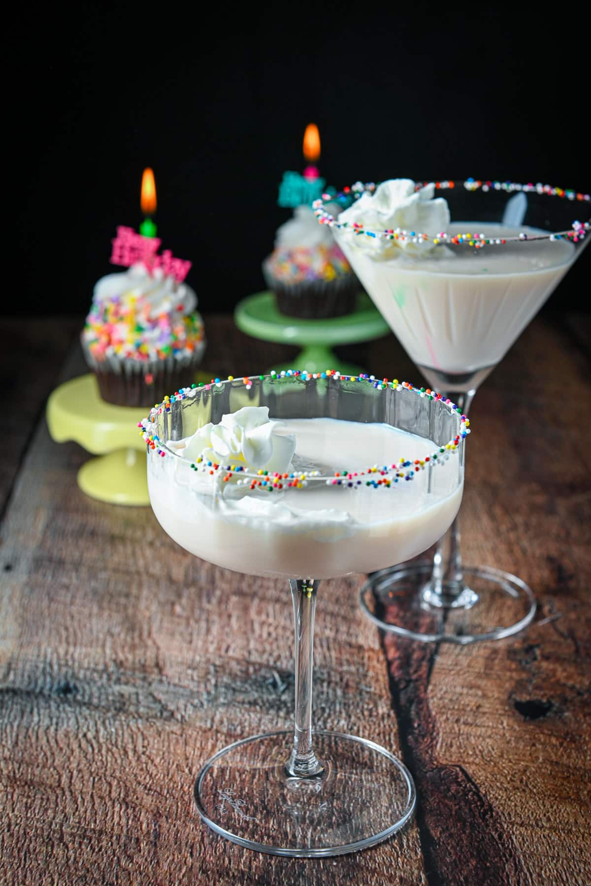 Two martini glasses filled with a cream drink with two cupcakes in the back