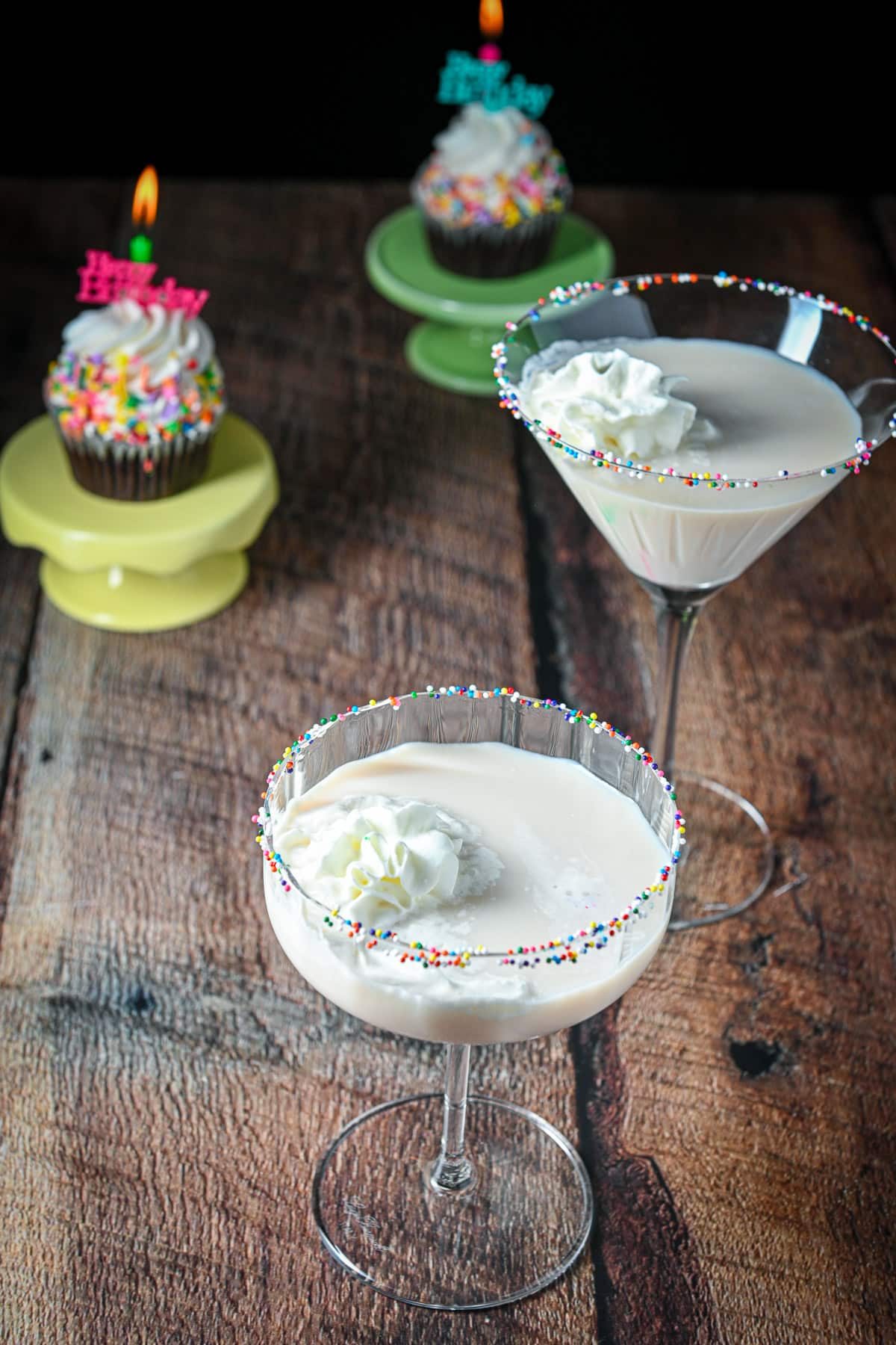 Two cupcakes on little pedestals behind two cocktails garnished with whipped cream and small colorful balls