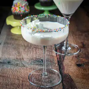 A creamy cocktail in a martini glass with colorful balls on the rim and cupcakes in the back
