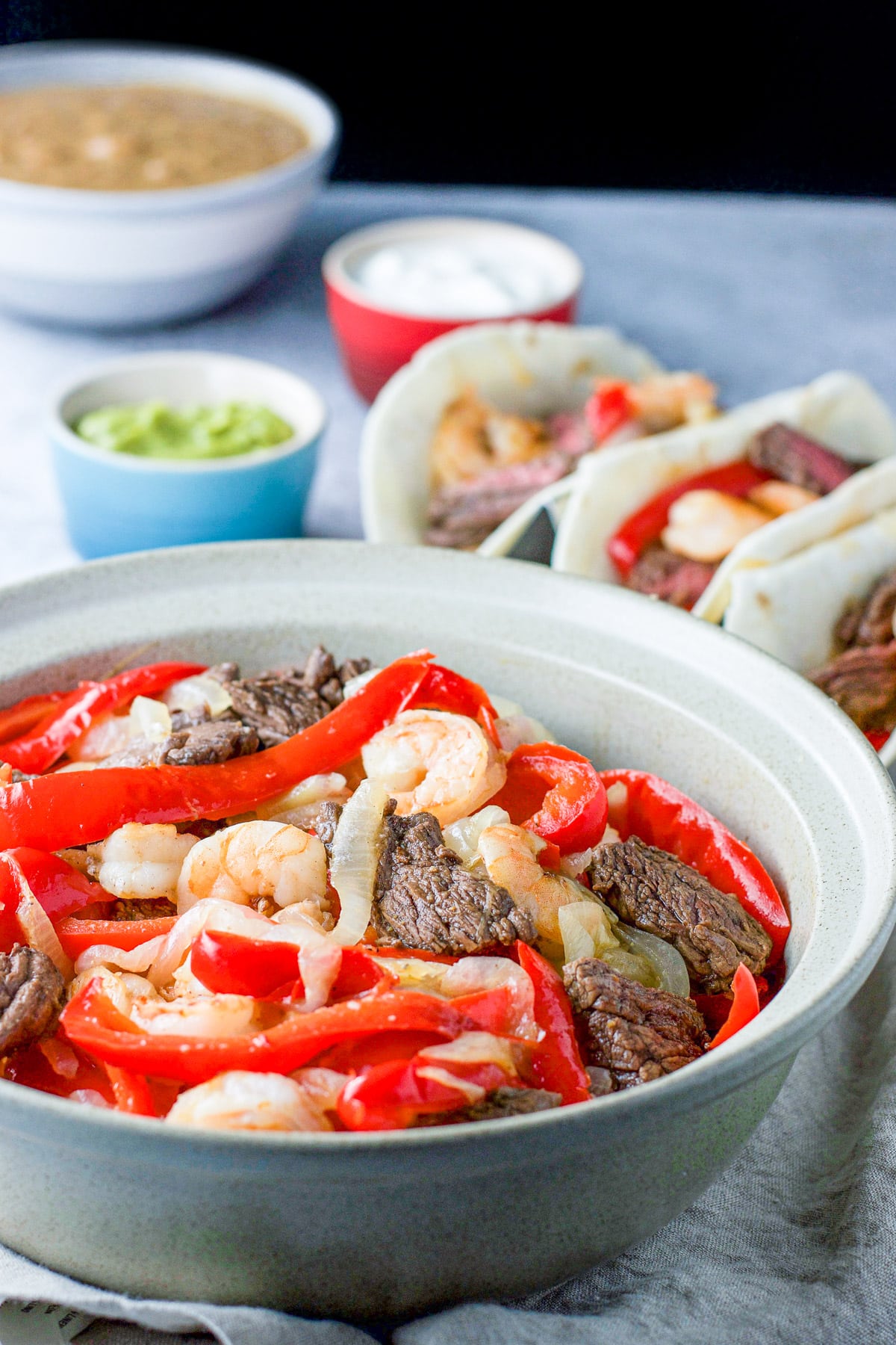 A bowl of shrimp and steak fajitas in front of some tacos, beans and garnish