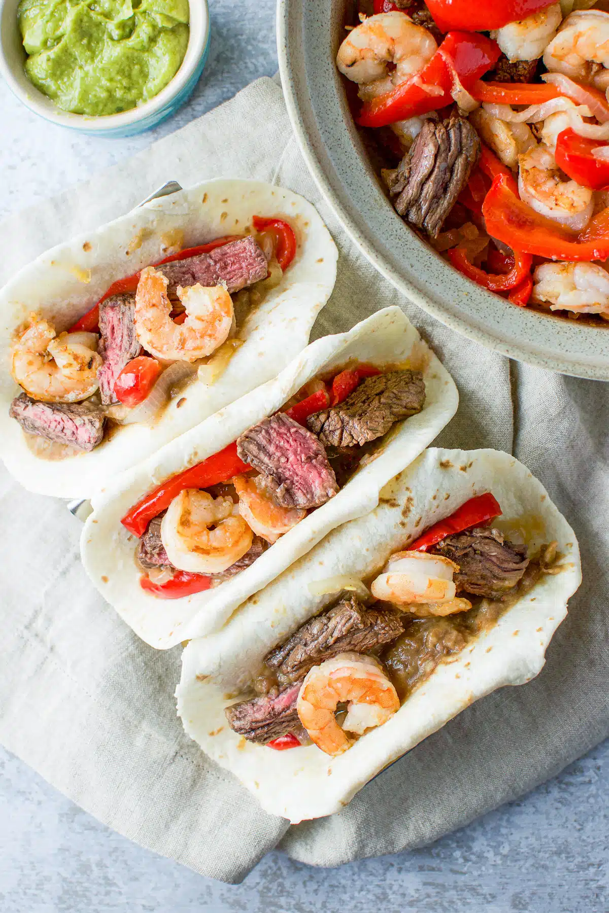 overhead view of three flour tortillas filled with fajitas and a bowl with the dish and guacamole