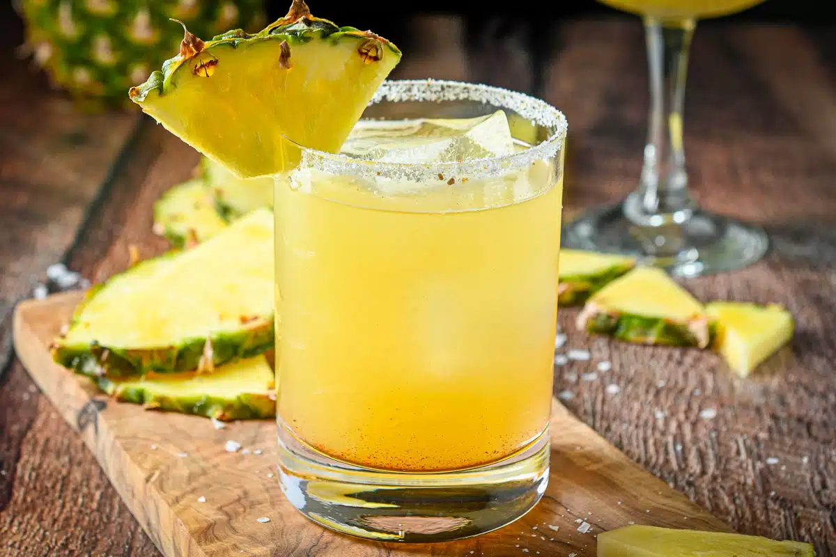horizontal photo of a salted glass with a yellow margarita in it with pineapple on the rim