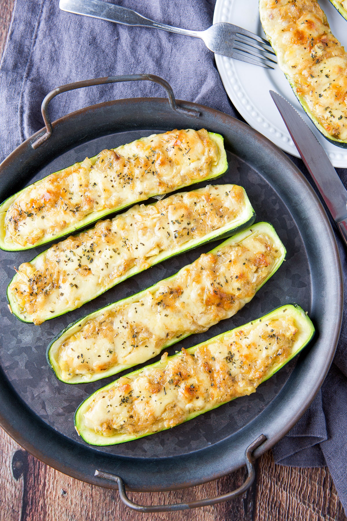 Overhead view of stuffed zucchini on a tray with a fork and knife on a plate with more of the vegetable