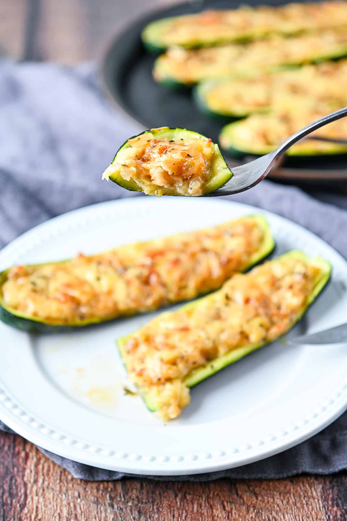 A fork with some stuffed zucchini on it held over a plate and a tray of zucchini behind