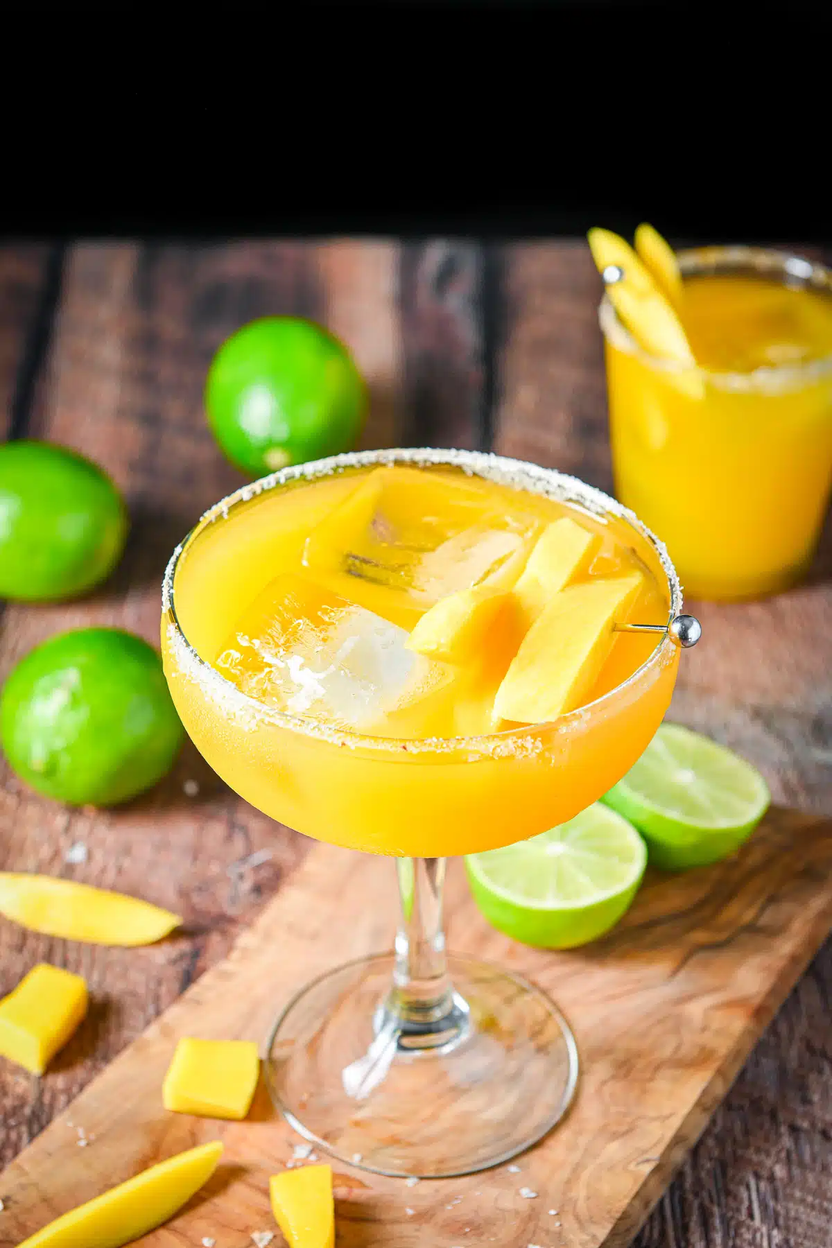 A classic margarita glass with the mango margarita on a board with slices of mango and limes