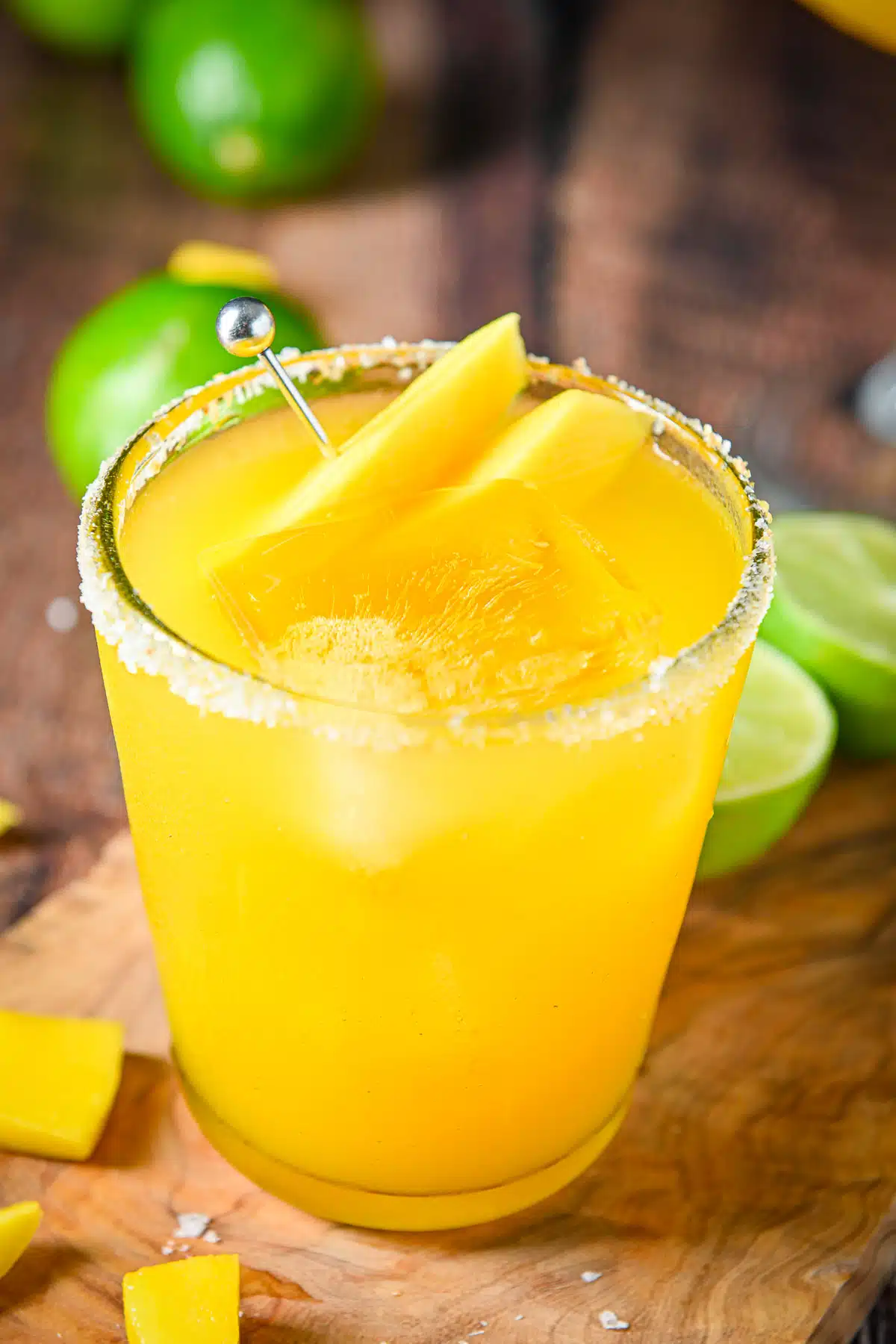 Close up of a margarita in a glass with mangos as garnish and limes on the table
