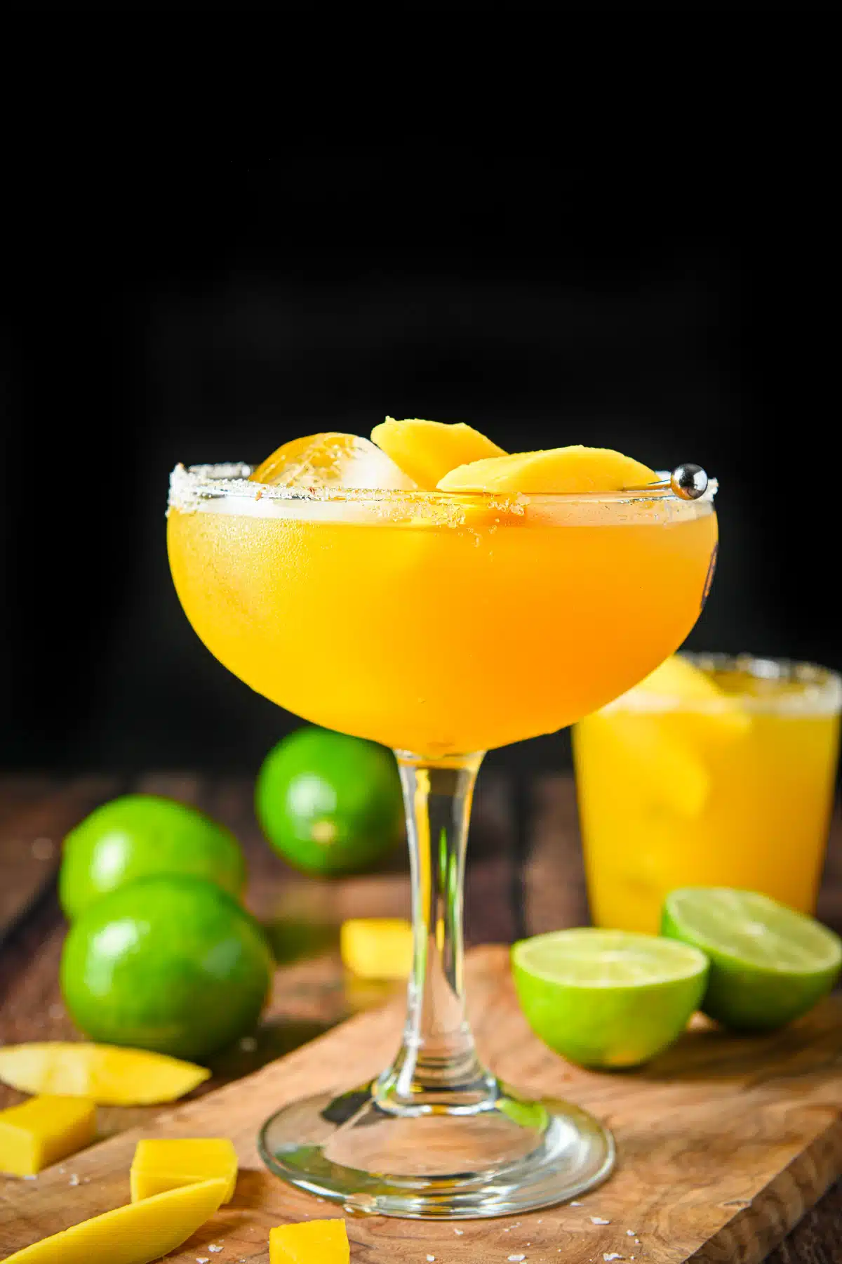 Vertical view of a bowl margarita glass filled with the mango drink with limes and mangos