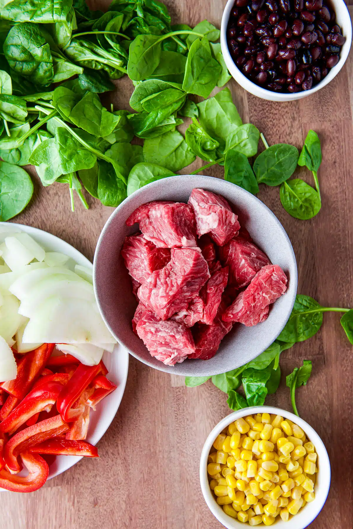 Overhead view of bowls filled with raw steak tips, corn, black beans, onion, and red bell peppers, and baby spinach