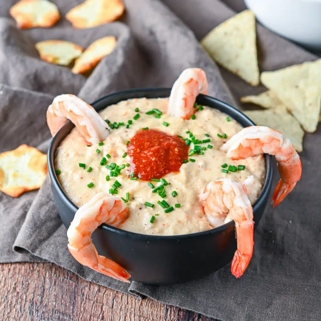 Square photo of a bowl filled with shrimp dip with shrimp on the rim and chips on the table