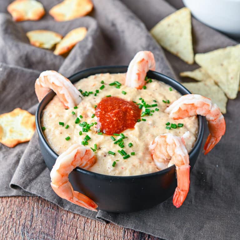 Square photo of a bowl filled with shrimp dip with shrimp on the rim and chips on the table