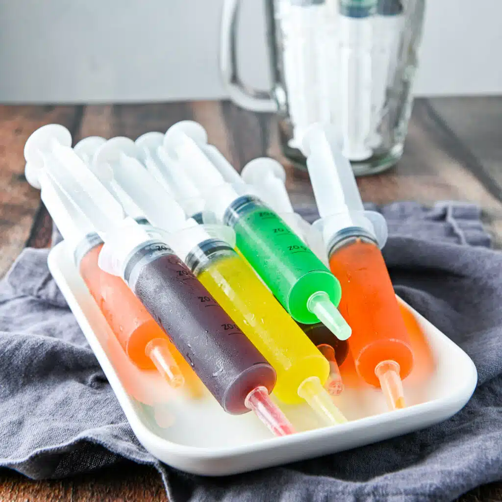 square photo of a white rectrangle plate with different colored jello shots in syringes