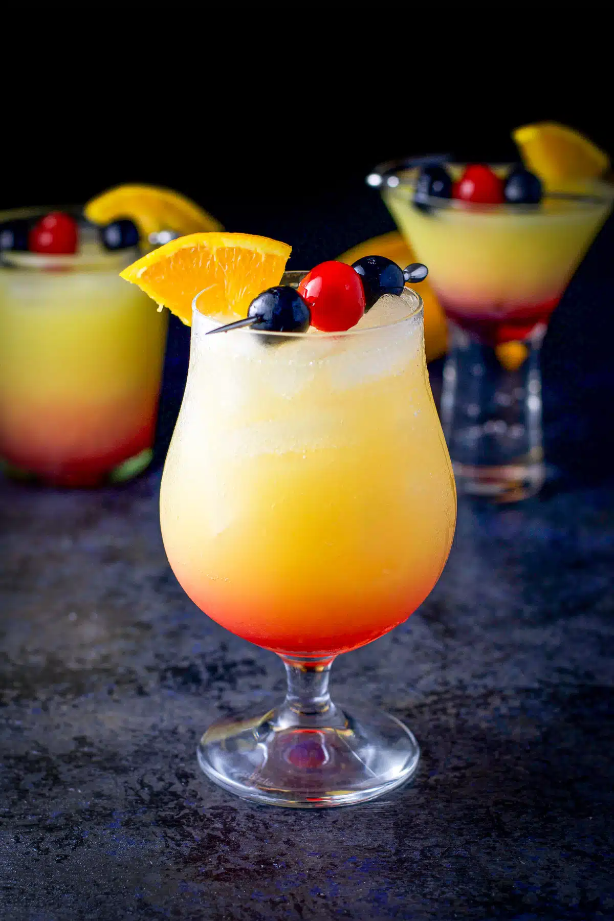 A tulip glass is in front of two other glasses filled with the tequila sunrise cocktail with cherries and orange wedges