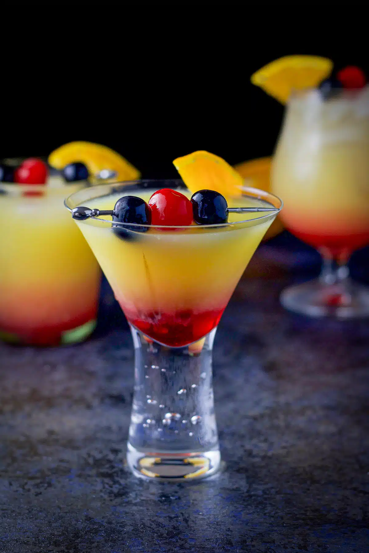 martini glass in front of two other shaped glasses filled with the orange/red cocktail with fruit garnish