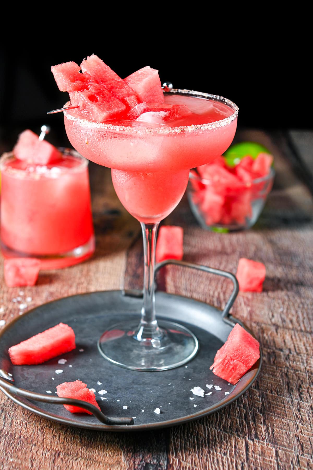 Tall classic margarita glass on the metal tray with the short glass in the back. There is watermelon on the tray, as garnish and in a little bowl