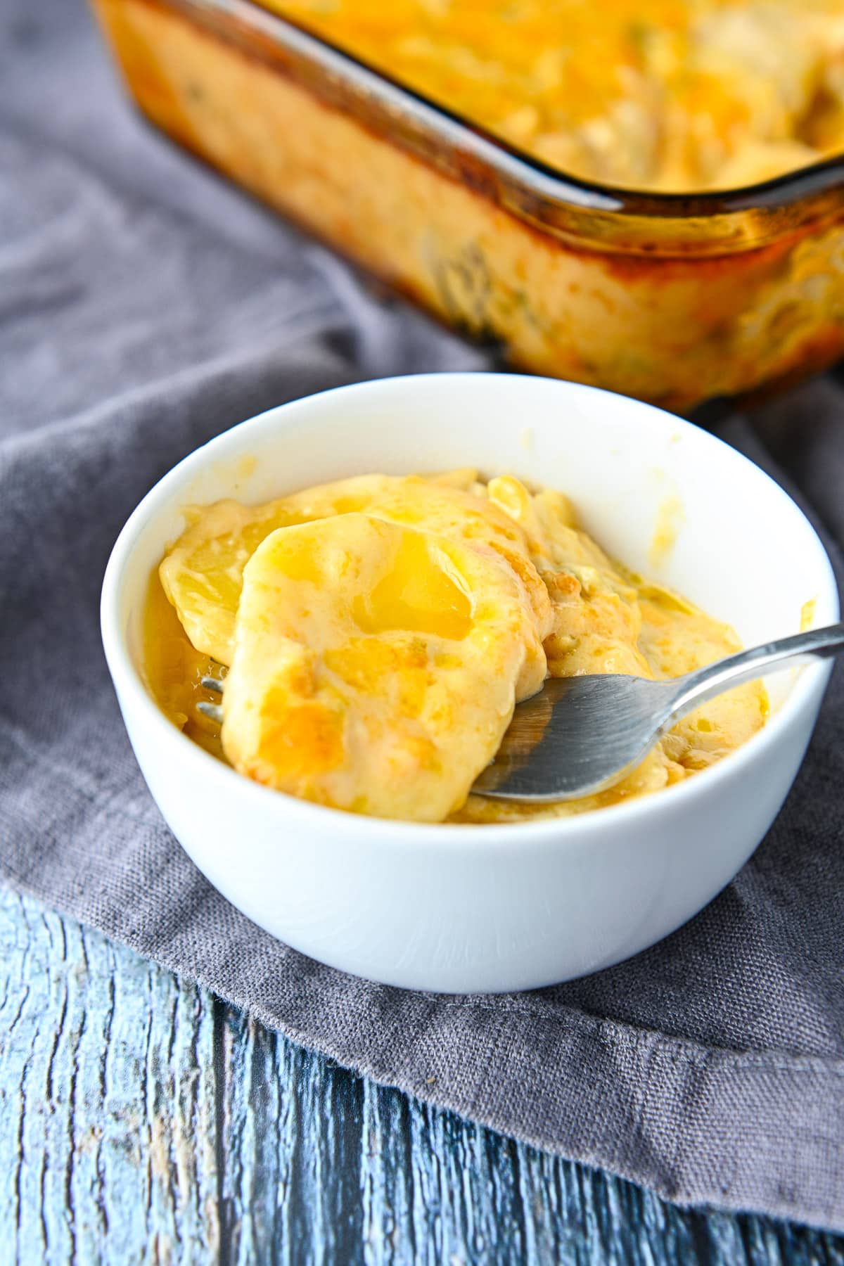 A small white bowl with the creamy potatoes in it along with a fork and the casserole in the back