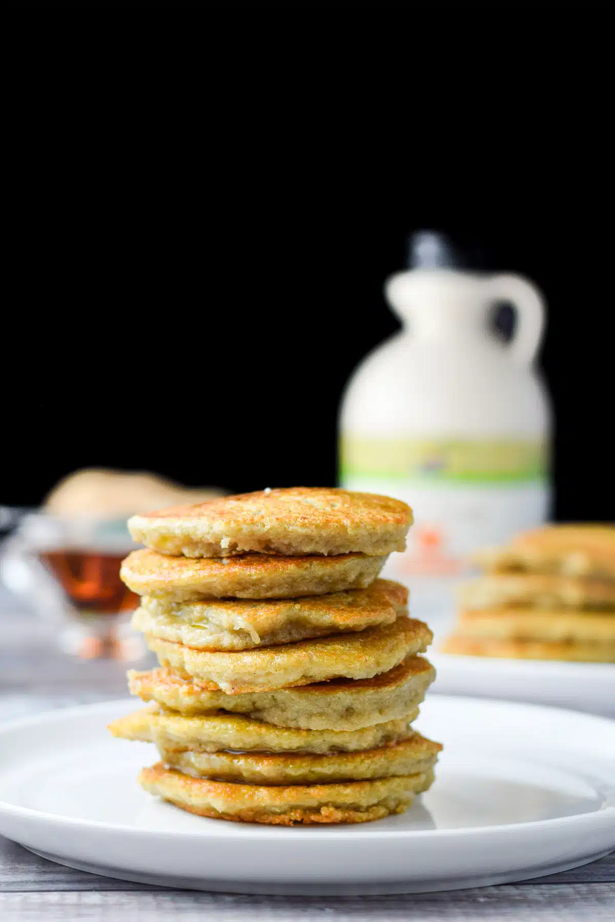 A white plate with a stack of pancakes and more behind along with syrup