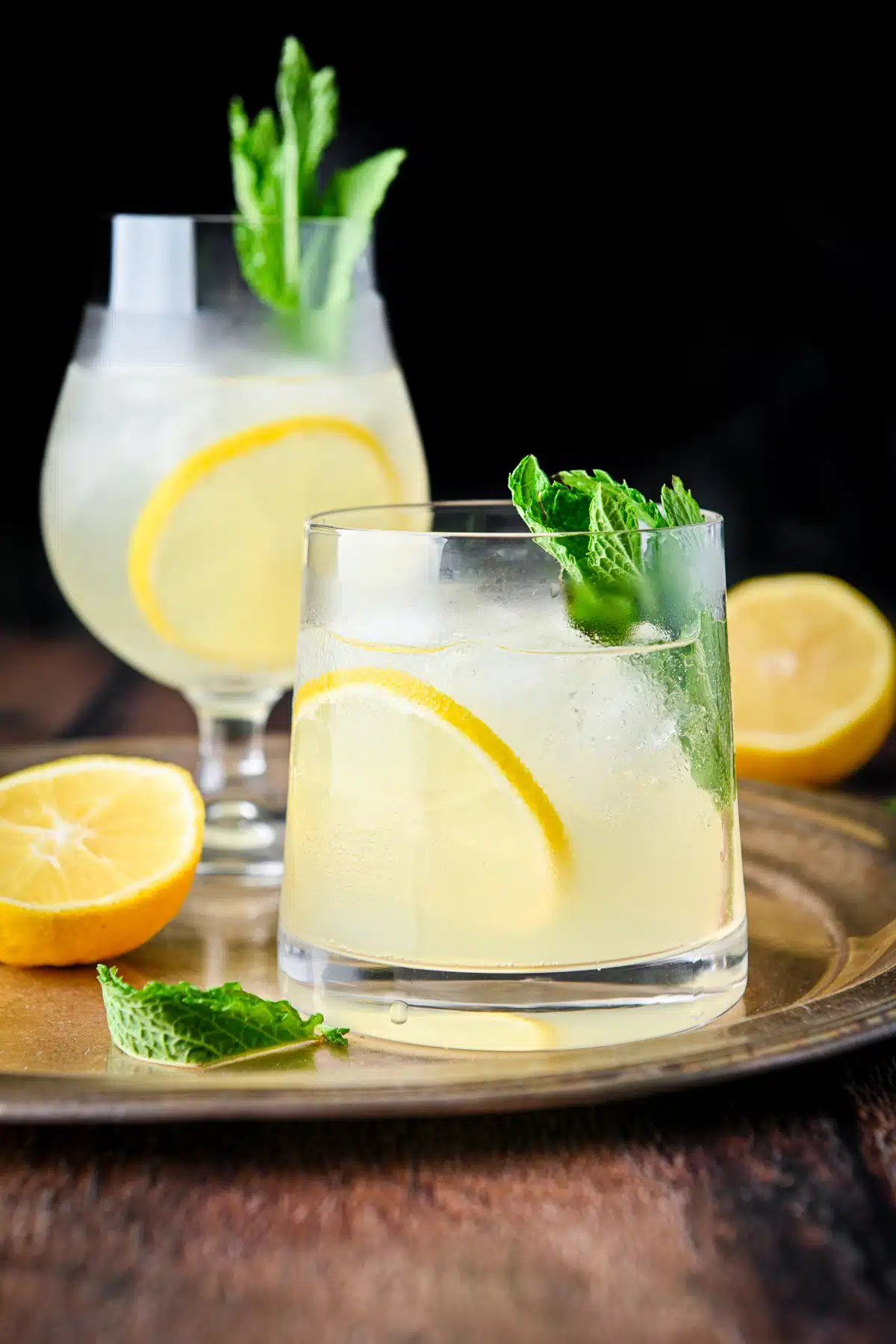 A double old-fashioned glass in front of a tulip shaped glass filled with the limoncello drink with lemon and mint