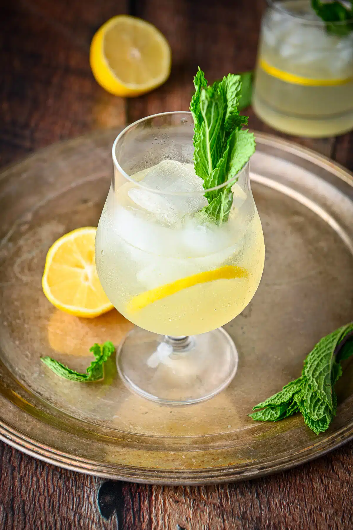 The tulip glass filled with the lemon cocktail on a metal tray with lemon slices and mint leaves