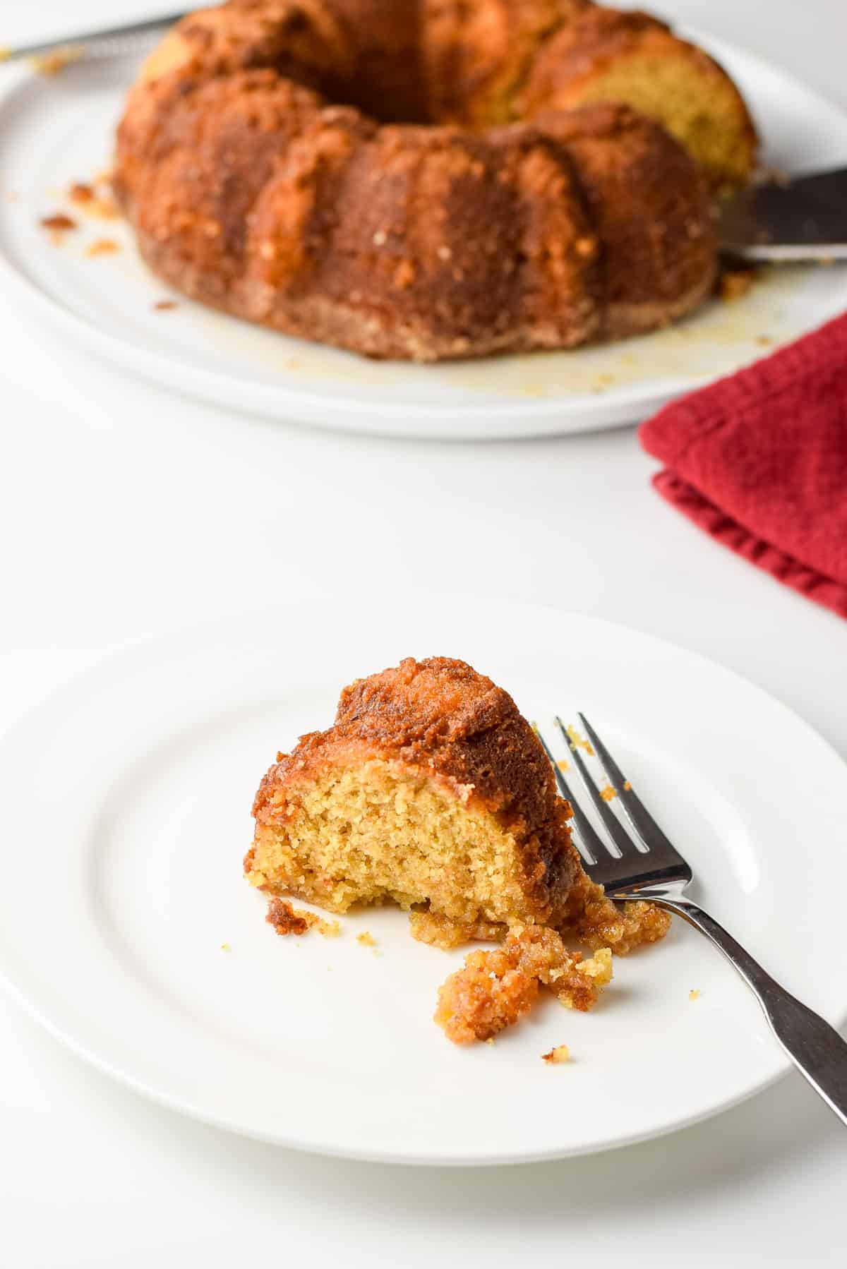 A little white plate with a slice of cake on it along with a fork and the cake on a big plate in the back