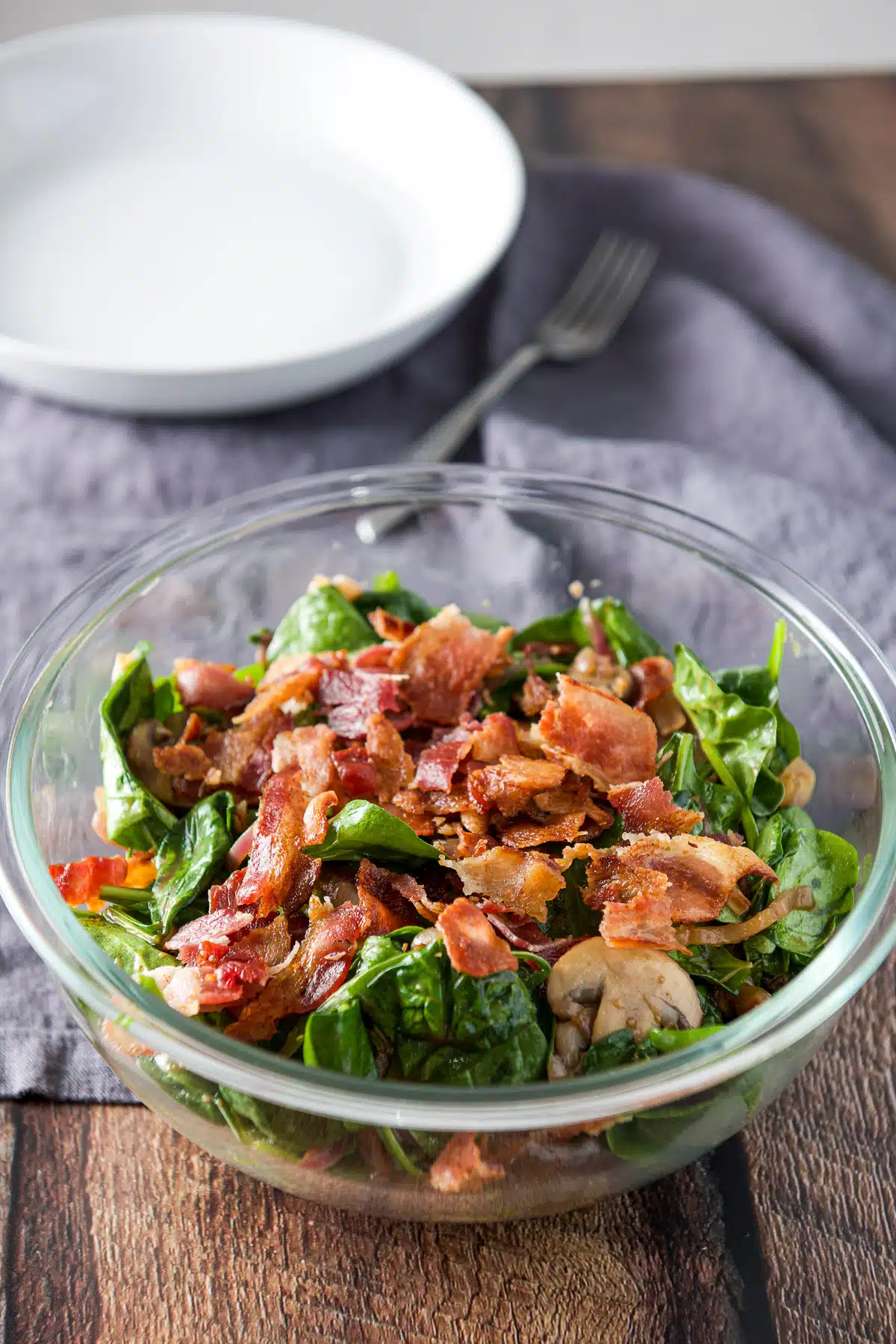 A glass bowl with the salad topped with pieces of bacon on a table with a napkin, fork and deep dish