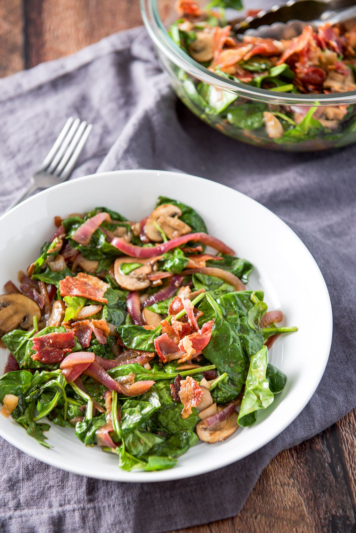 A glass bowl filled with a salad behind a white deep dish with the salad with mushrooms