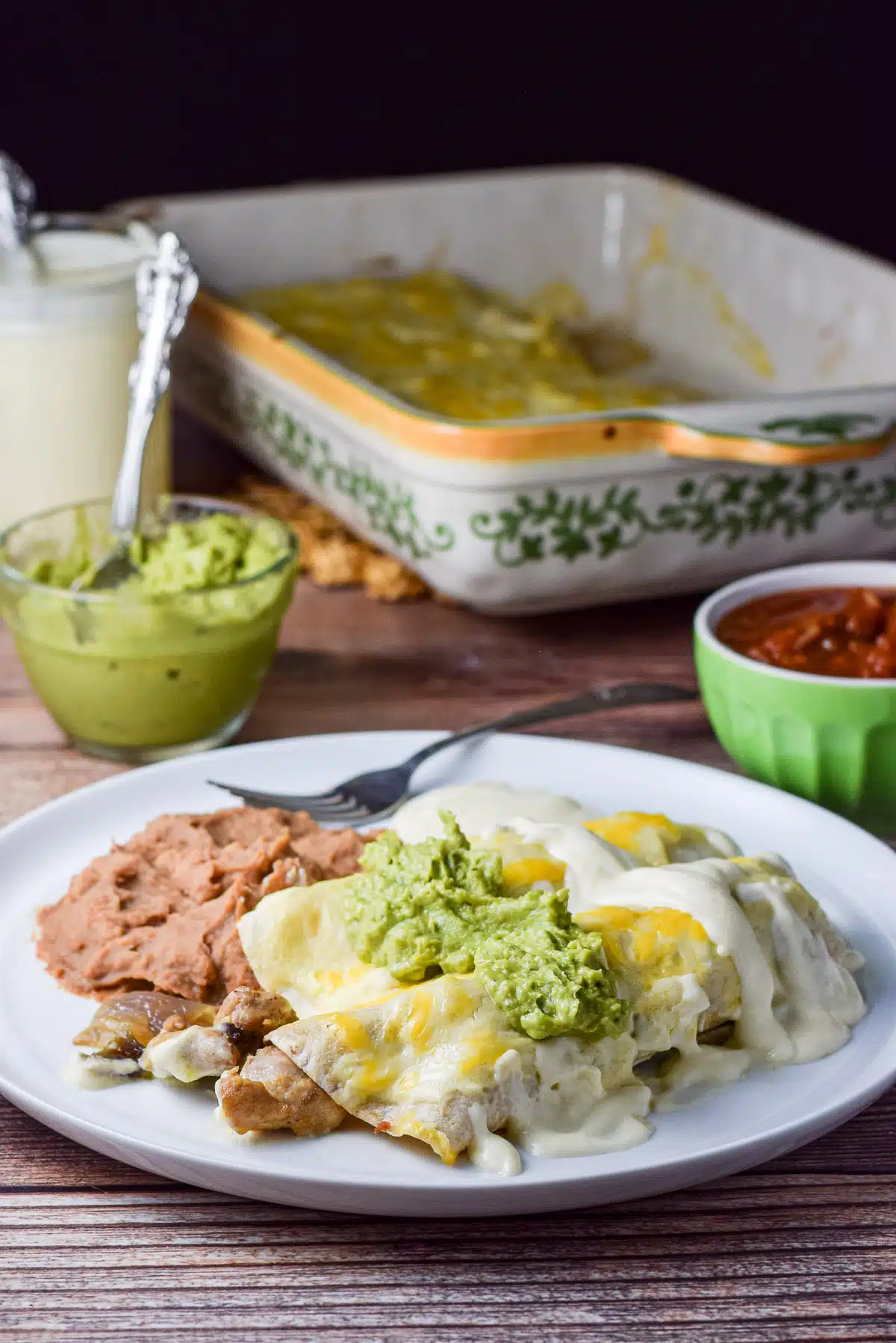 A white plate with chicken enchiladas with guacamole, and beans in front of guacamole, salsa, and sour cream sauce