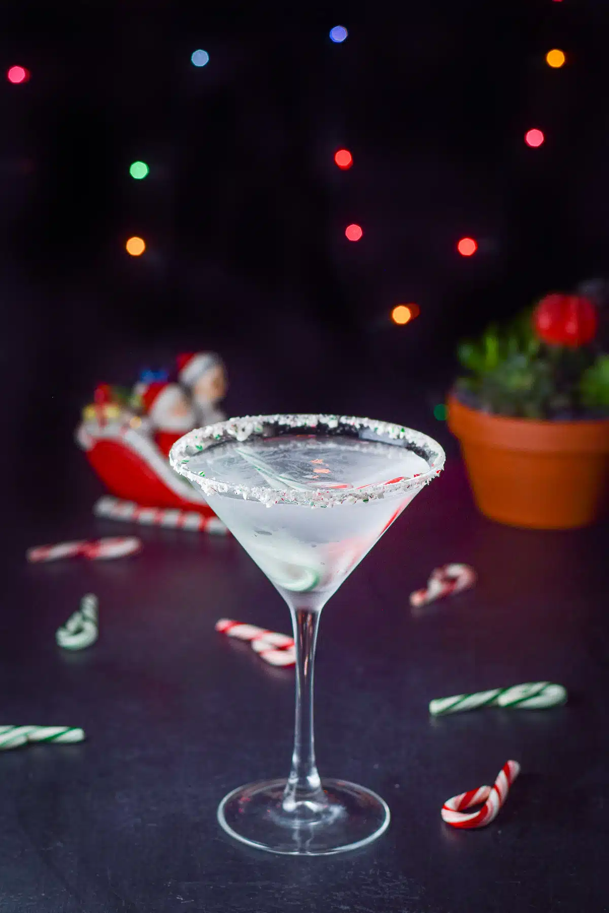 A glass with peppermint crushed on the rim is filled with a clear cocktail. There are lights in the back and candy canes on the table