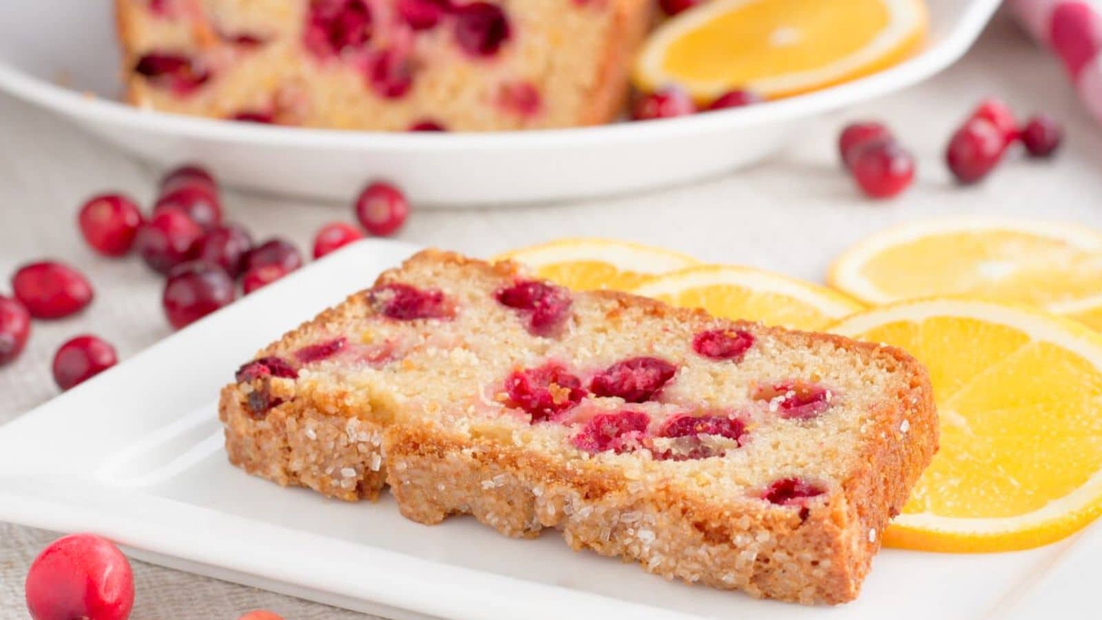 A white plate with a slice of cranberry bread on it with oranges and cranberries on the plate and table