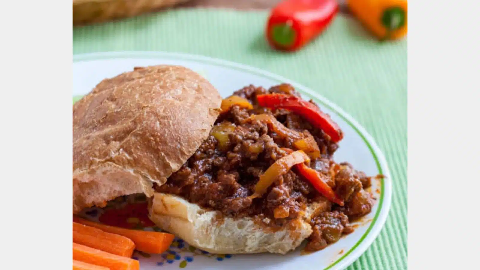 a plate with sloppy joes on a bun with carrots, and peppers in the back
