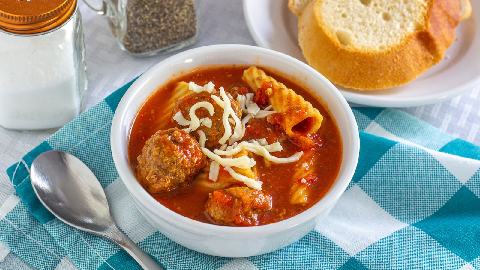 a bowl of meatball soup with macaroni and cheese on a napkin with bread, salt, and pepper in the back