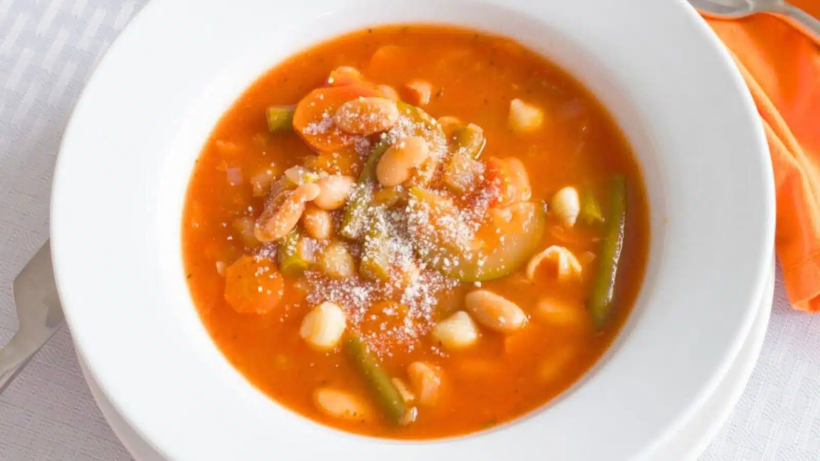 A white bowl filled with a tomato-based soup with vegetables, pasta and beans