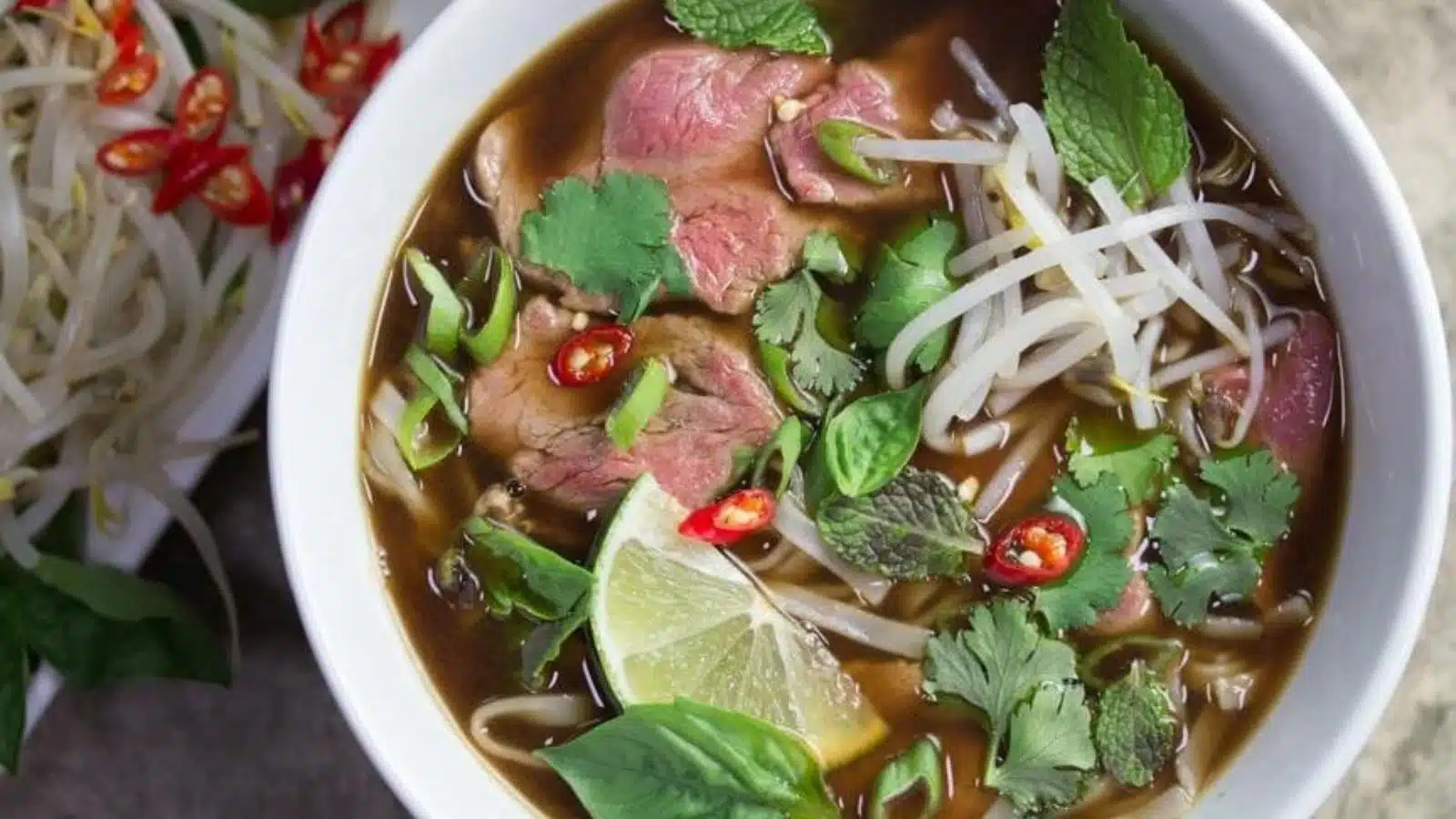 An overhead view of a white bowl with pho in it with beef, cilantro, mint, lime, and chiles