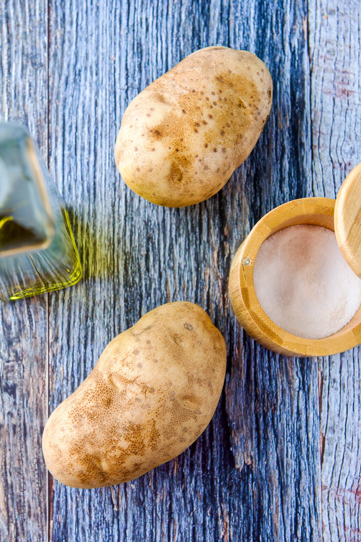Overhead view of two potatoes, salt, and oil on a blue wood table