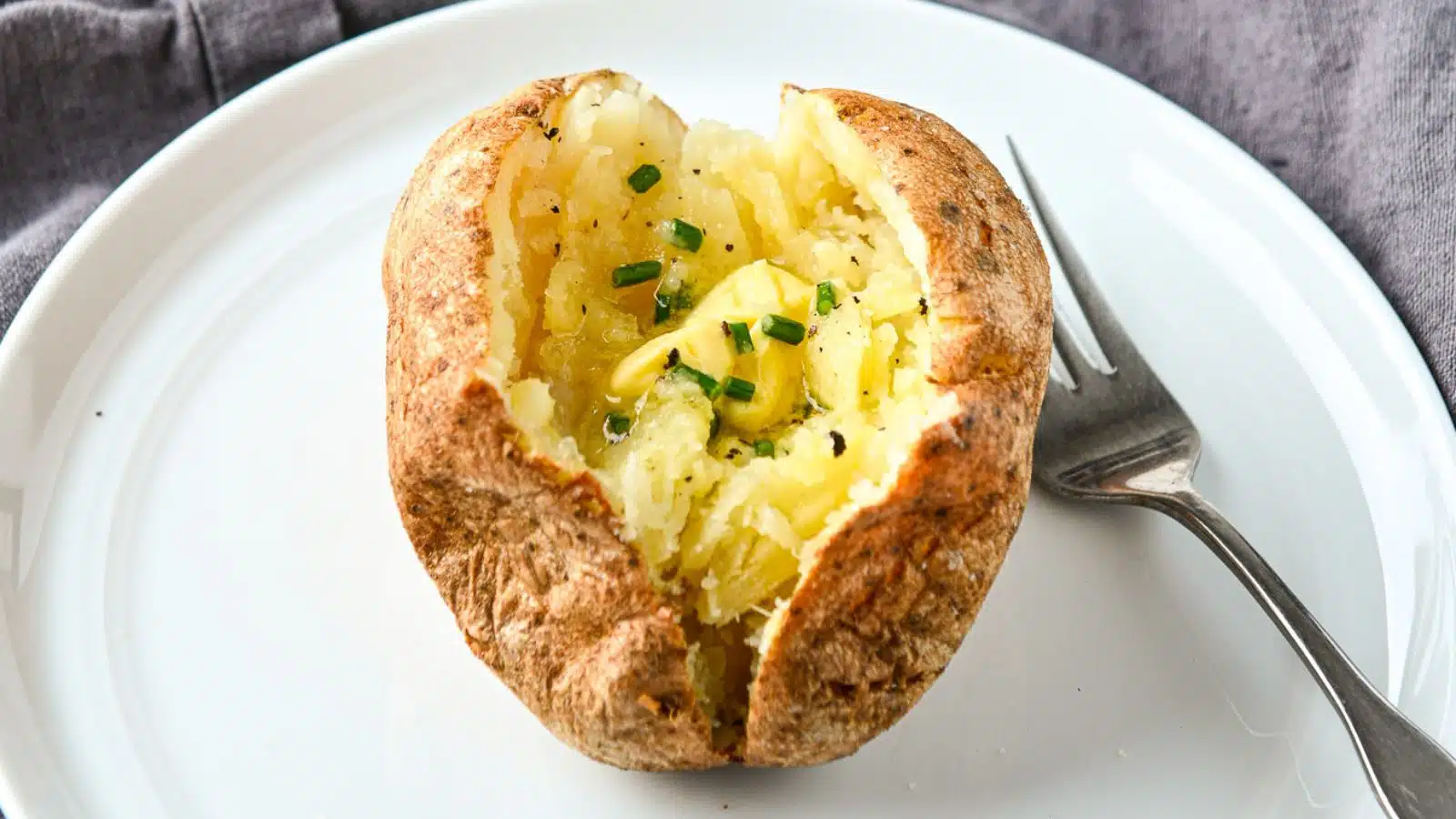a horizontal photo of a white plate with a fork and baked potato on it
