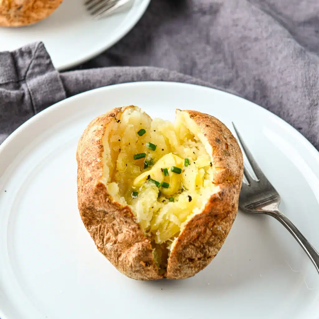a square photo of a white plate on a grey napkin with a baked potato on it with butter and chives
