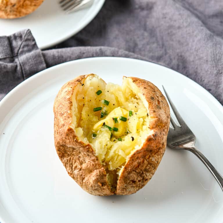 a square photo of a white plate on a grey napkin with a baked potato on it with butter and chives